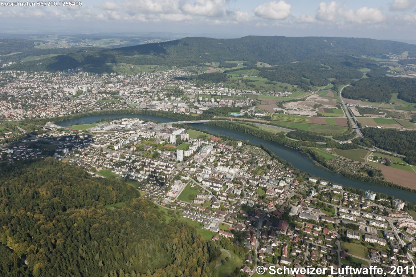 Wettingen - Neuenhof (Position: 2'667'284.47, 1'256'359.70). Im Bildzentrum Brücke der A1 Zürich-Bern über die Limmat. Ganz links im Bild: Kraftwerk 'Wettingen-Neuenhof' (Betrieb seit 1933) mit davor liegendem 'Stausee Neuenhof', einem gefragten Erholungsgebiet. - Im Vordergrund des Bildes (links der Limmat): Neuenhof; rechts der Limmat (Bildmitte links): Wettingen. Hügelzug im Hintergrund: Lägern